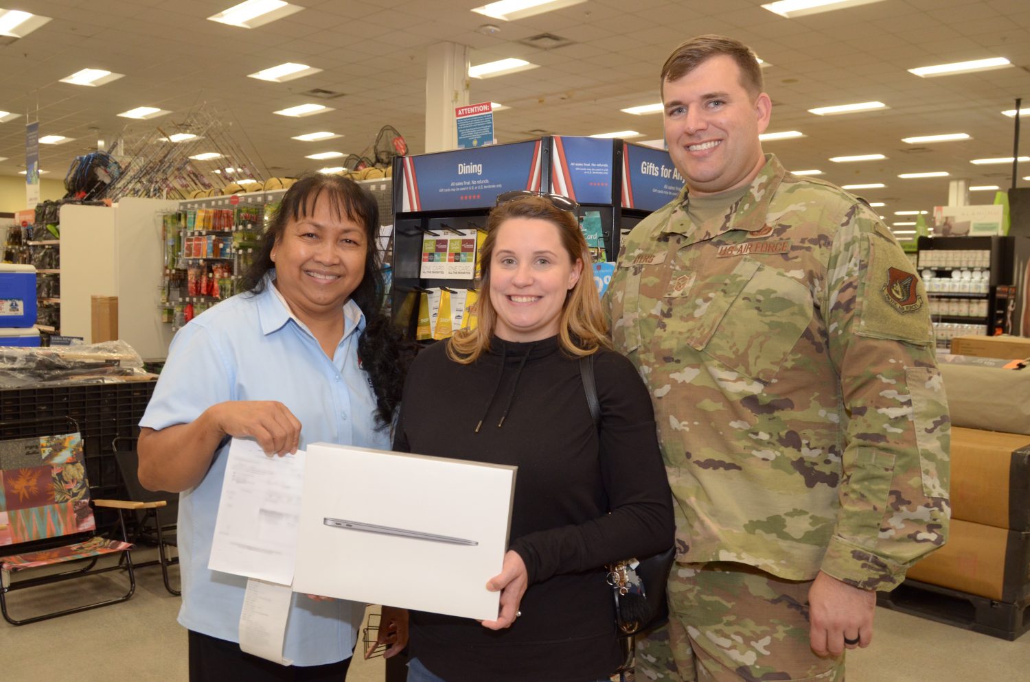 Pay Away the Layaway Dec PAC event 1 <stromg>Air Force Master Sgt. Brett Myers, Engineering Craftsman with the 718th Civil Engineer Squadron, and his wife, Misti, pose for a picture with Flor Payton, Kadena Exchange main store manager, after Brett surprised Misti with a MacBook Pro. Brett used the Exchange’s layaway program for the first time to purchase the laptop for Misti, who is pursuing a degree in medical administration. On Dec. 15, Brett was invited to a video call with celebrity and Marine and Navy Veteran, Montel Williams. During the call, Williams announced that all Exchange layaways in the Pacific Region, including Brett’s, were paid in full by nonprofit Pay Away the Layaway. (U.S. Army Photo by Staff Sgt. Mark A. Kauffman)</strong>