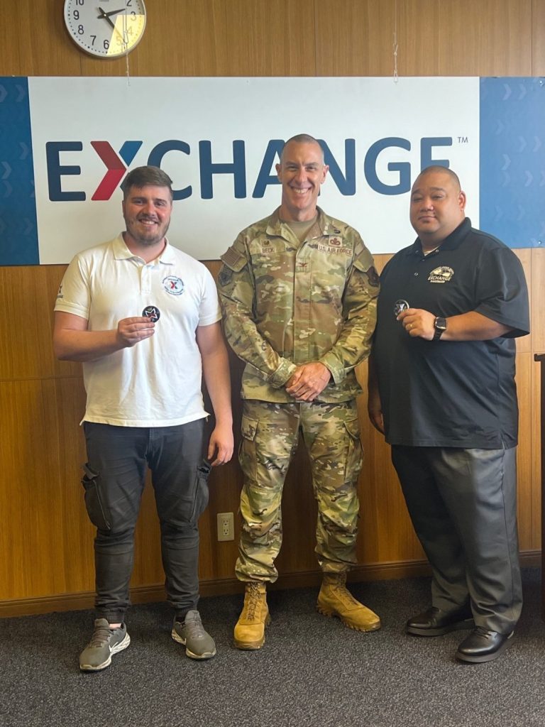 Repair & Maintenance Supervisor Dimitrios Diamantopoulos, Pacific Region Commander Col. Jason Beck and Food Plant Operations Manager Uji Battle in front of Exchange sign.