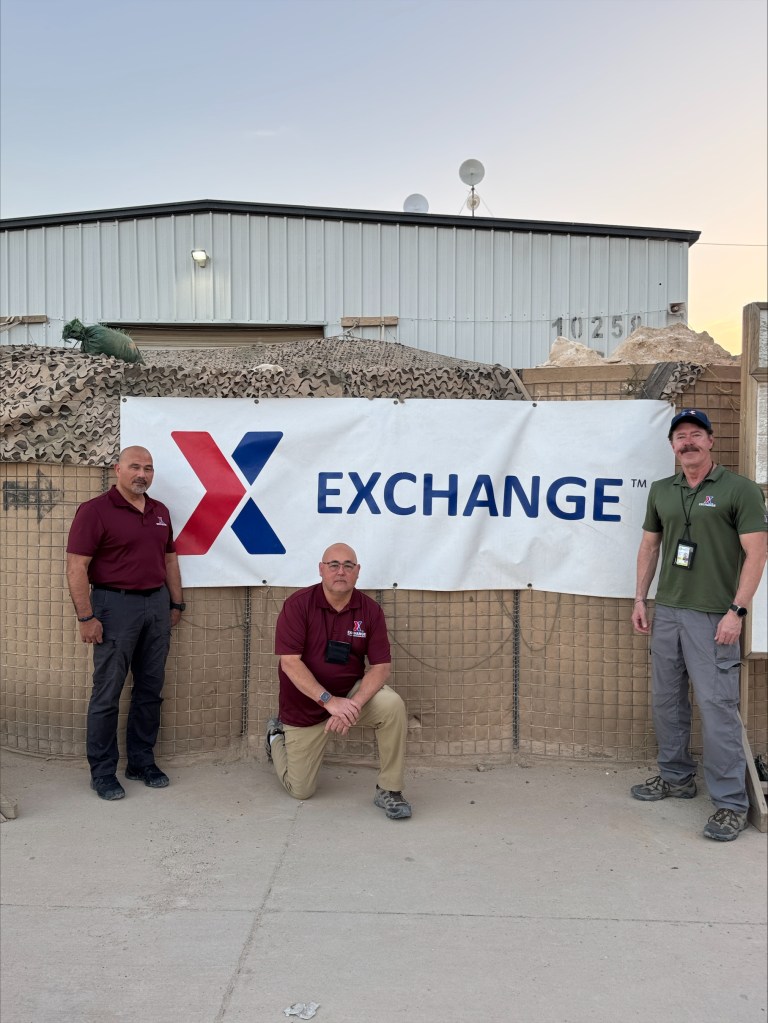 Three men in front of an Exchange banner in Iraq.